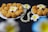 A close-up view of two plates filled with golden-brown fried snacks, garnished with white frangipani flowers. A strand of pearly beads is elegantly placed beside the plates on a dark textured background.