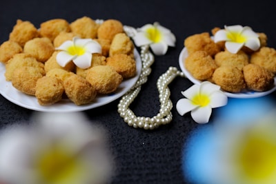 A close-up view of two plates filled with golden-brown fried snacks, garnished with white frangipani flowers. A strand of pearly beads is elegantly placed beside the plates on a dark textured background.