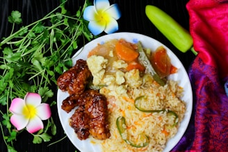 A white plate with a vibrant meal consisting of fried rice garnished with green chilies, stir-fried vegetables including carrots and okra, and glazed crispy meat. Fresh cilantro leaves, a green chili, and decorative flowers are placed around the plate. A colorful red and purple fabric adds to the aesthetic of the composition.