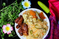 A white plate with a vibrant meal consisting of fried rice garnished with green chilies, stir-fried vegetables including carrots and okra, and glazed crispy meat. Fresh cilantro leaves, a green chili, and decorative flowers are placed around the plate. A colorful red and purple fabric adds to the aesthetic of the composition.