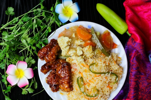 A white plate with a vibrant meal consisting of fried rice garnished with green chilies, stir-fried vegetables including carrots and okra, and glazed crispy meat. Fresh cilantro leaves, a green chili, and decorative flowers are placed around the plate. A colorful red and purple fabric adds to the aesthetic of the composition.