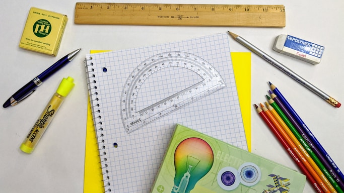A variety of stationery items arranged on a surface, including a spiral notebook with graph paper, a wooden ruler, a protractor, a mechanical pencil, colored pencils, a yellow highlighter, a pen, an eraser, and a small box. The colors are vibrant and the items are neatly organized.