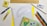 A variety of stationery items arranged on a surface, including a spiral notebook with graph paper, a wooden ruler, a protractor, a mechanical pencil, colored pencils, a yellow highlighter, a pen, an eraser, and a small box. The colors are vibrant and the items are neatly organized.