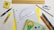 A variety of stationery items arranged on a surface, including a spiral notebook with graph paper, a wooden ruler, a protractor, a mechanical pencil, colored pencils, a yellow highlighter, a pen, an eraser, and a small box. The colors are vibrant and the items are neatly organized.