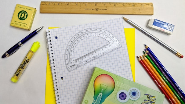 A variety of stationery items arranged on a surface, including a spiral notebook with graph paper, a wooden ruler, a protractor, a mechanical pencil, colored pencils, a yellow highlighter, a pen, an eraser, and a small box. The colors are vibrant and the items are neatly organized.