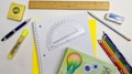 A variety of stationery items arranged on a surface, including a spiral notebook with graph paper, a wooden ruler, a protractor, a mechanical pencil, colored pencils, a yellow highlighter, a pen, an eraser, and a small box. The colors are vibrant and the items are neatly organized.