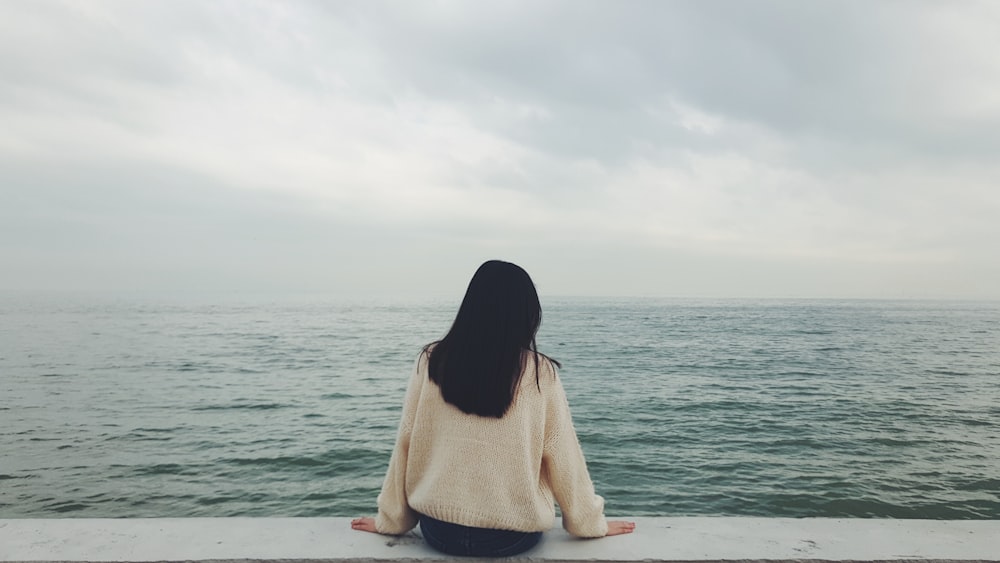 woman in white sweater sitting on white sand near body of water during daytime