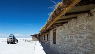 A rustic building with walls made of bricks and a roof covered in straw stands in a vast, flat landscape. A vehicle with baggage on its roof is parked on the white ground, suggesting a remote or desert-like environment under a clear, bright blue sky.