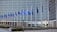 Several blue flags with yellow stars are arranged on tall flagpoles in front of a large, modern office building with a grid-like glass facade. The flags are gently waving and are representative of the European Union.
