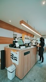A modern coffee shop's counter area with a barista standing behind it. The counter is made of wood and equipped with various coffee machines and a digital display. Several paper pockets are attached to the side, and a trash bin is placed at the corner. The ceiling features recessed lighting and a long, linear pendant light runs over the counter.