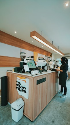 A modern coffee shop's counter area with a barista standing behind it. The counter is made of wood and equipped with various coffee machines and a digital display. Several paper pockets are attached to the side, and a trash bin is placed at the corner. The ceiling features recessed lighting and a long, linear pendant light runs over the counter.
