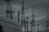 A group of tall transmission towers and power lines dominate the scene under a cloudy, overcast sky. A white van passes by in the foreground, partially visible. The atmosphere is dark and moody, suggesting an impending storm.