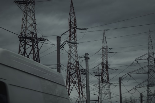 A group of tall transmission towers and power lines dominate the scene under a cloudy, overcast sky. A white van passes by in the foreground, partially visible. The atmosphere is dark and moody, suggesting an impending storm.