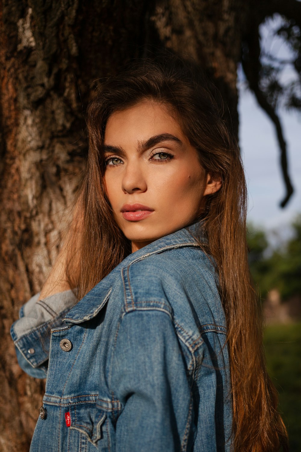 woman in blue denim jacket leaning on brown tree during daytime