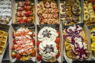 An assortment of decorated waffles arranged in boxes. Each waffle is topped with various fruits like strawberries, bananas, and kiwi, as well as toppings such as whipped cream, chocolate, and sprinkles. The vibrant colors and intricate details of the toppings create an appetizing display.