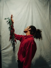 A person with curly hair stands sideways while wearing a red shirt. They are holding a green leafy vine with red ribbons near their raised hand. The backdrop is a plain, light-colored cloth.