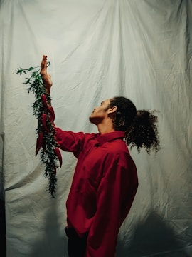 A person with curly hair stands sideways while wearing a red shirt. They are holding a green leafy vine with red ribbons near their raised hand. The backdrop is a plain, light-colored cloth.