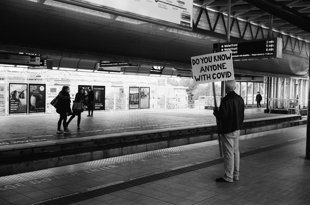 grayscale photo of man in black jacket and white pants standing on train station
