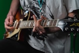 A person wearing a white graphic T-shirt plays an electric guitar with a sunburst finish. The focus is on their hands and the guitar, specifically on the strings and tuning pegs. The background is a solid green color.