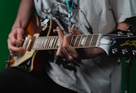 A person wearing a white graphic T-shirt plays an electric guitar with a sunburst finish. The focus is on their hands and the guitar, specifically on the strings and tuning pegs. The background is a solid green color.
