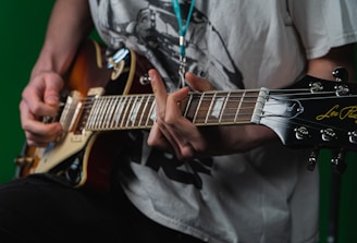 A person wearing a white graphic T-shirt plays an electric guitar with a sunburst finish. The focus is on their hands and the guitar, specifically on the strings and tuning pegs. The background is a solid green color.
