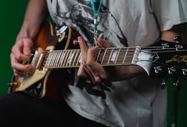 A person wearing a white graphic T-shirt plays an electric guitar with a sunburst finish. The focus is on their hands and the guitar, specifically on the strings and tuning pegs. The background is a solid green color.