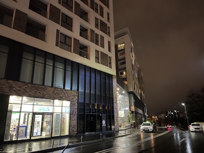 A modern multi-story building at night with a lit retail store front displaying advertisements in the window. The building features tall windows and a combination of brick and concrete materials. Several cars are parked on the street, which is wet from rain, reflecting the city lights. The sky is cloudy, contributing to the dim ambiance.