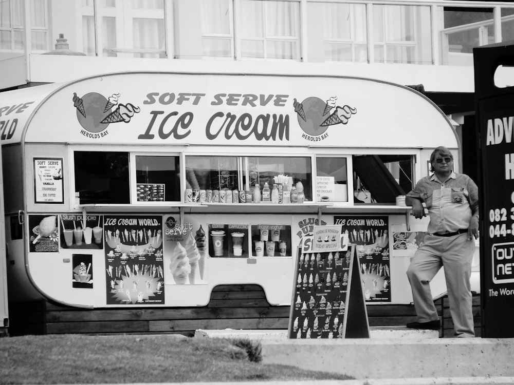 grayscale photo of man in black jacket and pants standing beside food cart