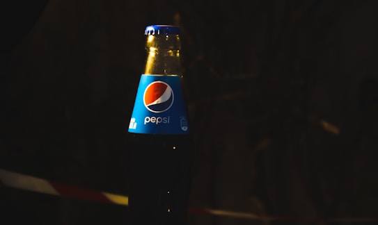 A glass bottle of Pepsi with a blue label is positioned against a dark background. The label prominently displays the Pepsi logo, with a curved red, white, and blue circle above the Pepsi name in white letters. The bottle is illuminated by focused light, creating a contrast against the shadowy backdrop.