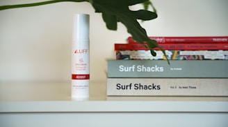 A white bottle with the label 'LUFF CBD Cream Assist' is placed next to a stack of books. The books have titles related to 'Surf Shacks'. A large, dark green leaf partially covers the top of the books.