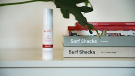 A white bottle with the label 'LUFF CBD Cream Assist' is placed next to a stack of books. The books have titles related to 'Surf Shacks'. A large, dark green leaf partially covers the top of the books.