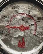 A close-up of a thermometer gauge, partially obscured by ice or frost, creating a textured and cold appearance. The dial numbers and red pointer are visible beneath the icy surface, giving a sense of freezing temperatures.