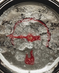 A close-up of a thermometer gauge, partially obscured by ice or frost, creating a textured and cold appearance. The dial numbers and red pointer are visible beneath the icy surface, giving a sense of freezing temperatures.