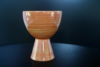 A ceramic piece with a unique, double-cone shape. The upper part is bowl-like, smoothly transitioning into a narrower base. The surface features horizontal, swirling orange and cream stripes, giving it a sleek and polished appearance. The background is a dark, monochrome surface that contrasts with the object's warm tones.