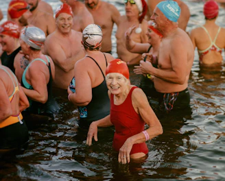 A group of swimmers wearing colorful swim caps and various swimsuits stand together in a body of water. The atmosphere appears lively and joyful, with people smiling and interacting with one another.