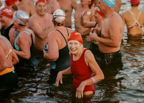 A group of swimmers wearing colorful swim caps and various swimsuits stand together in a body of water. The atmosphere appears lively and joyful, with people smiling and interacting with one another.