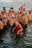 A group of swimmers wearing colorful swim caps and various swimsuits stand together in a body of water. The atmosphere appears lively and joyful, with people smiling and interacting with one another.