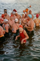 A group of swimmers wearing colorful swim caps and various swimsuits stand together in a body of water. The atmosphere appears lively and joyful, with people smiling and interacting with one another.