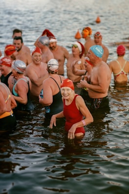 A group of swimmers wearing colorful swim caps and various swimsuits stand together in a body of water. The atmosphere appears lively and joyful, with people smiling and interacting with one another.