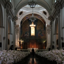 A beautifully adorned interior of a church with a high arched ceiling and elaborate chandeliers. The walls display detailed frescoes with religious imagery. Rows of white flowers line the aisle, leading to an altar featuring a prominent cross and religious statues.