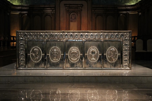 An ornate, metallic altar with intricate carvings and embossed religious figures arranged in circular medallions. It is set against a dark, wooden backdrop with high, arched panels.