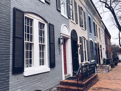 A row of charming, historic townhouses with brick facades and various colored exteriors, featuring black shutters and doors. Each house has distinct architectural details such as arched windows and ornate lanterns. The sidewalk is made of bricks, and barren trees line the quiet street, creating an atmospheric urban setting.
