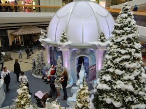 A festive shopping mall scene featuring a large holiday display with a winter theme. A white dome structure surrounded by artificial snow-covered Christmas trees dominates the setting. There are people interacting and taking photos, with one person dressed in a Santa Claus costume sitting with children. Nearby, adults are walking around, and a stroller is visible.