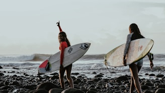 woman in red bikini holding white surfboard standing on rocky shore during daytime