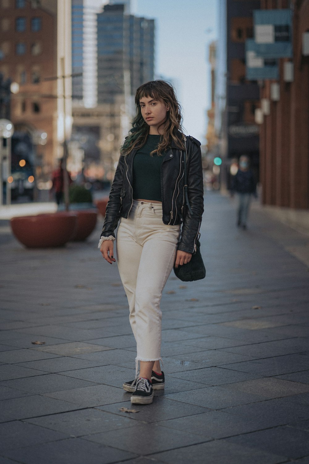 woman in black leather jacket and white pants standing on sidewalk during daytime