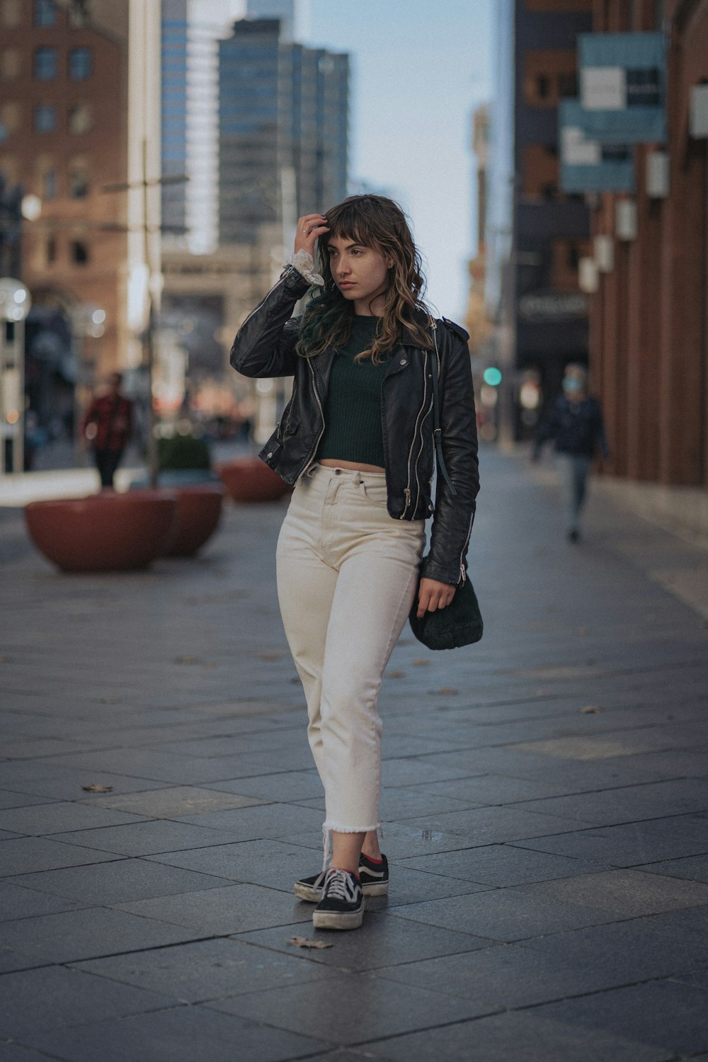 woman in black leather jacket and white pants standing on sidewalk during daytime