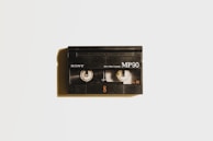 A vintage black Sony MP90 8mm video cassette is centered against a light background. It has visible spools through a transparent window and displays the text 'Sony', 'MP90', and 'PAL 8'. A subtle shadow casts to the left, adding depth.