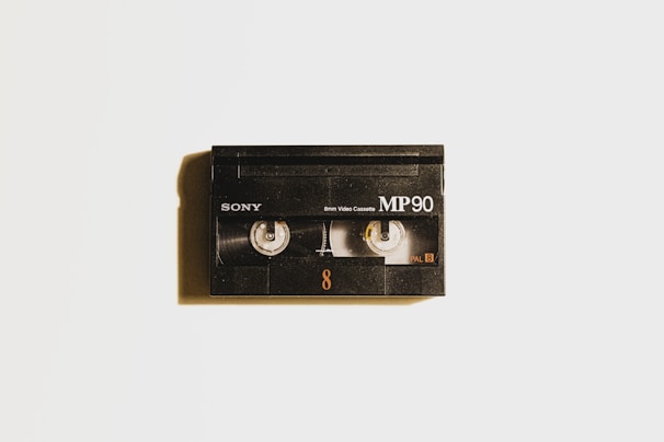 A vintage black Sony MP90 8mm video cassette is centered against a light background. It has visible spools through a transparent window and displays the text 'Sony', 'MP90', and 'PAL 8'. A subtle shadow casts to the left, adding depth.