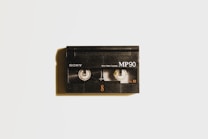 A vintage black Sony MP90 8mm video cassette is centered against a light background. It has visible spools through a transparent window and displays the text 'Sony', 'MP90', and 'PAL 8'. A subtle shadow casts to the left, adding depth.
