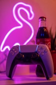 A gaming controller is placed on a surface with a dimly lit neon flamingo sign in the background. Next to the controller is a bottle of Coca-Cola, adding a retro vibe to the composition. The scene is surrounded by a soft pink and purple glow, creating a cozy and nostalgic atmosphere.